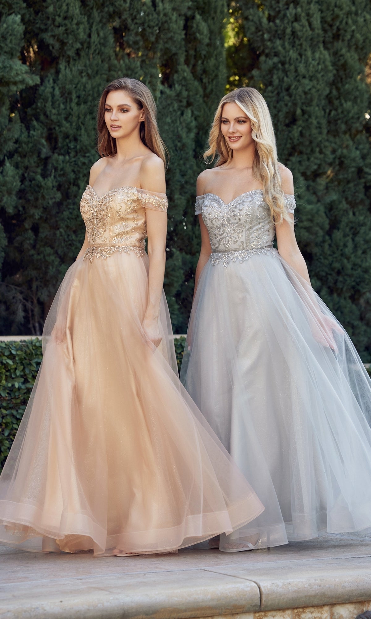 Two women in off-the-shoulder prom dresses stand outdoors; one wears the Long Sweetheart Prom Ball Gown: Juliet 287 in peach, while the other is in a silver dress with a beaded corset bodice, set against lush green trees.